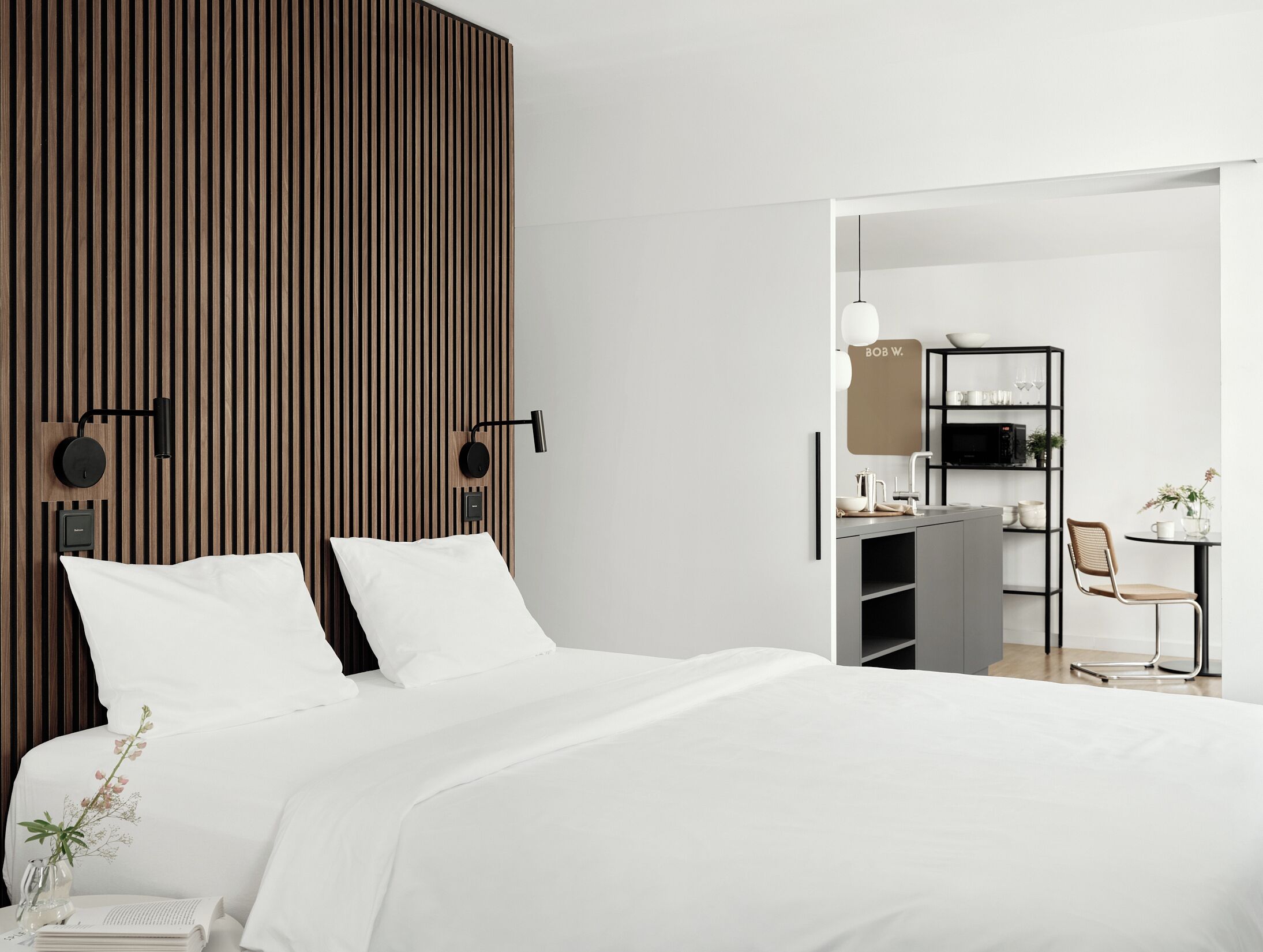 A modern bedroom with a bed, a wooden slat wall, two wall-mounted lights, a bedside table with a vase, and a view into a connected kitchen area.