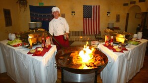 Un chef cocina en un wok grande con fuego, rodeado de mesas con comida y condimentos.