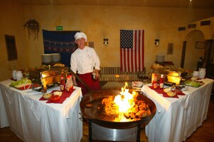 Un chef cocina en un wok grande con fuego, rodeado de mesas con comida y condimentos.