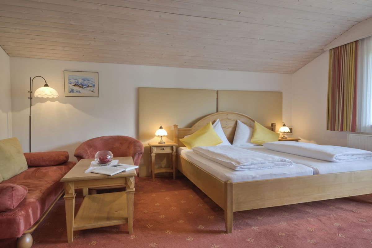 A hotel room with a bed, a wooden headboard, a small table with a lamp, a red sofa, and a framed picture on the wall.