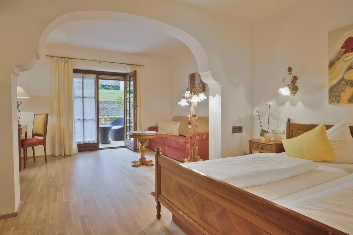 A bedroom with a wooden bed, a red sofa, a small table, and a view of the outdoors through a glass door.