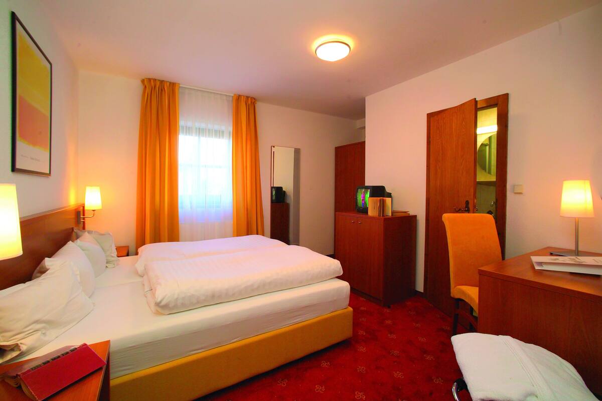 A hotel room with a double bed, a desk with a chair, a wardrobe, a mirror, and a red carpet.