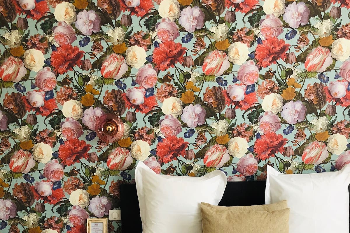 Habitación de hotel con papel tapiz floral, una cama con ropa de cama blanca y una mesita de noche con una lámpara y un certificado enmarcado.