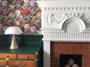 A room with a green chest of drawers, a white fireplace with a brick surround, and a floral wallpaper. - AMA Hôtel (Biarritz)