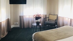 A hotel room with a bed, a chair, a small table with a vase, a flat-screen TV mounted on the wall, and a window with sheer curtains. - AMA Hôtel (Biarritz)