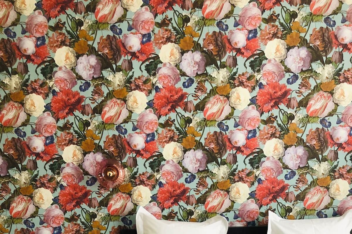 Una habitación de hotel con papel tapiz floral, una cama con sábanas blancas, un plaid beige y una mesita de noche con un teléfono y una foto enmarcada.