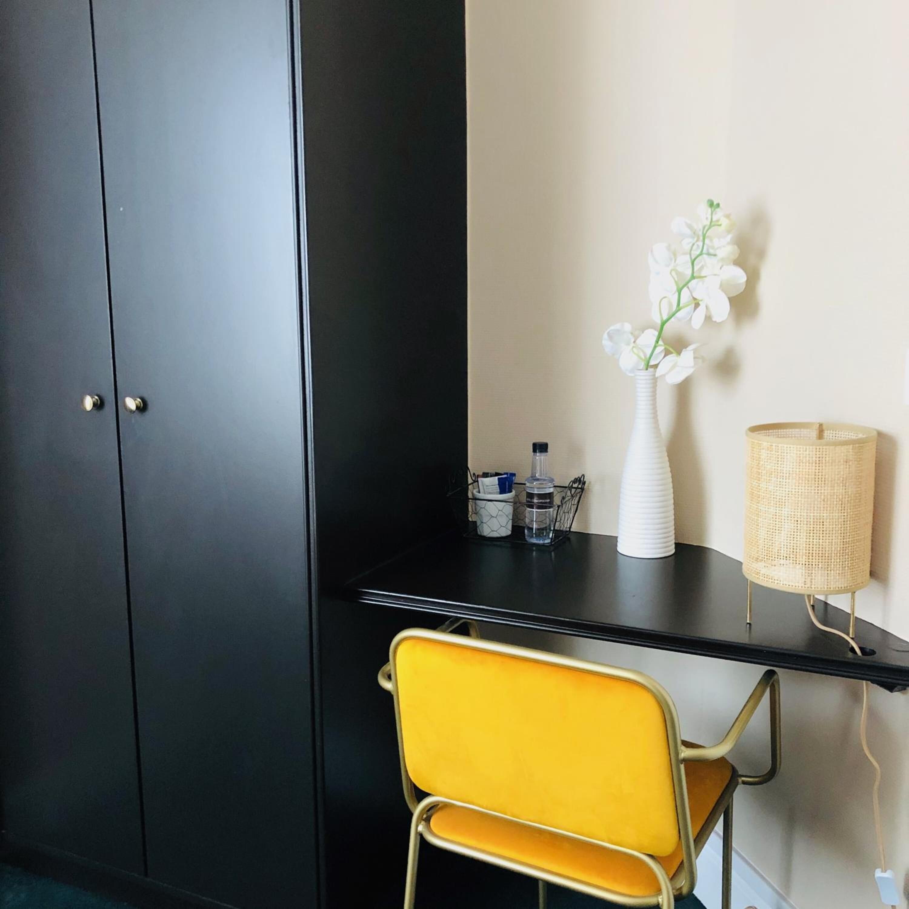 Um quarto com um guarda-roupa escuro, uma escrivaninha preta, uma cadeira amarela, um vaso branco com flores e uma luminária de tecido.
