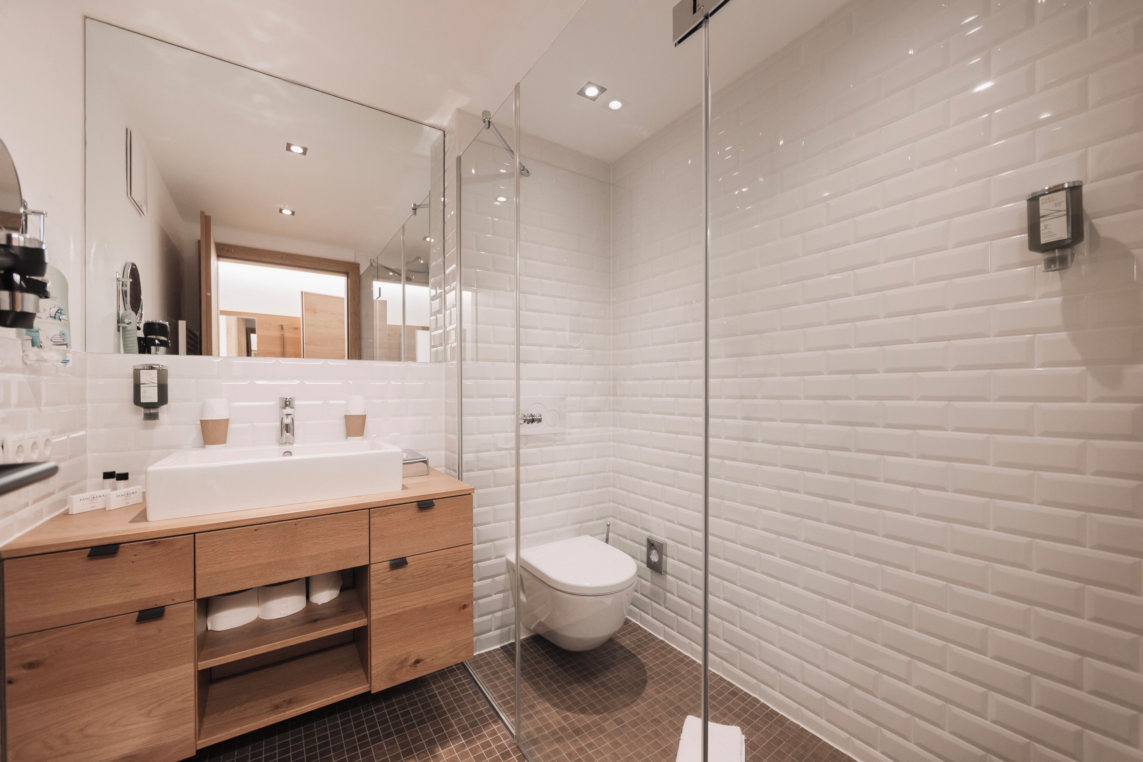 A modern bathroom with a large mirror, a wooden vanity, a white sink, and a glass-enclosed shower.