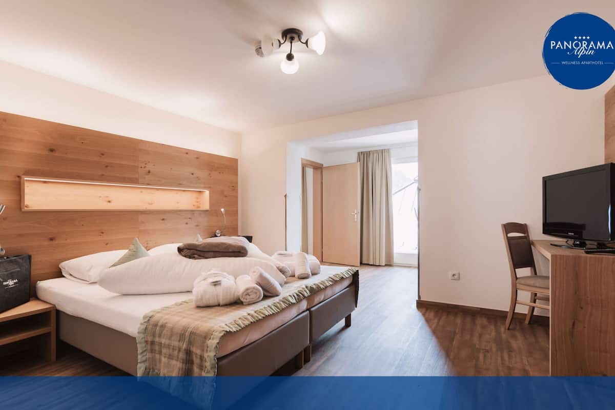 Une chambre d’hôtel avec une tête de lit en bois, un lit avec du linge de maison blanc, une table de chevet avec une lampe, un bureau avec une télévision et une chaise.