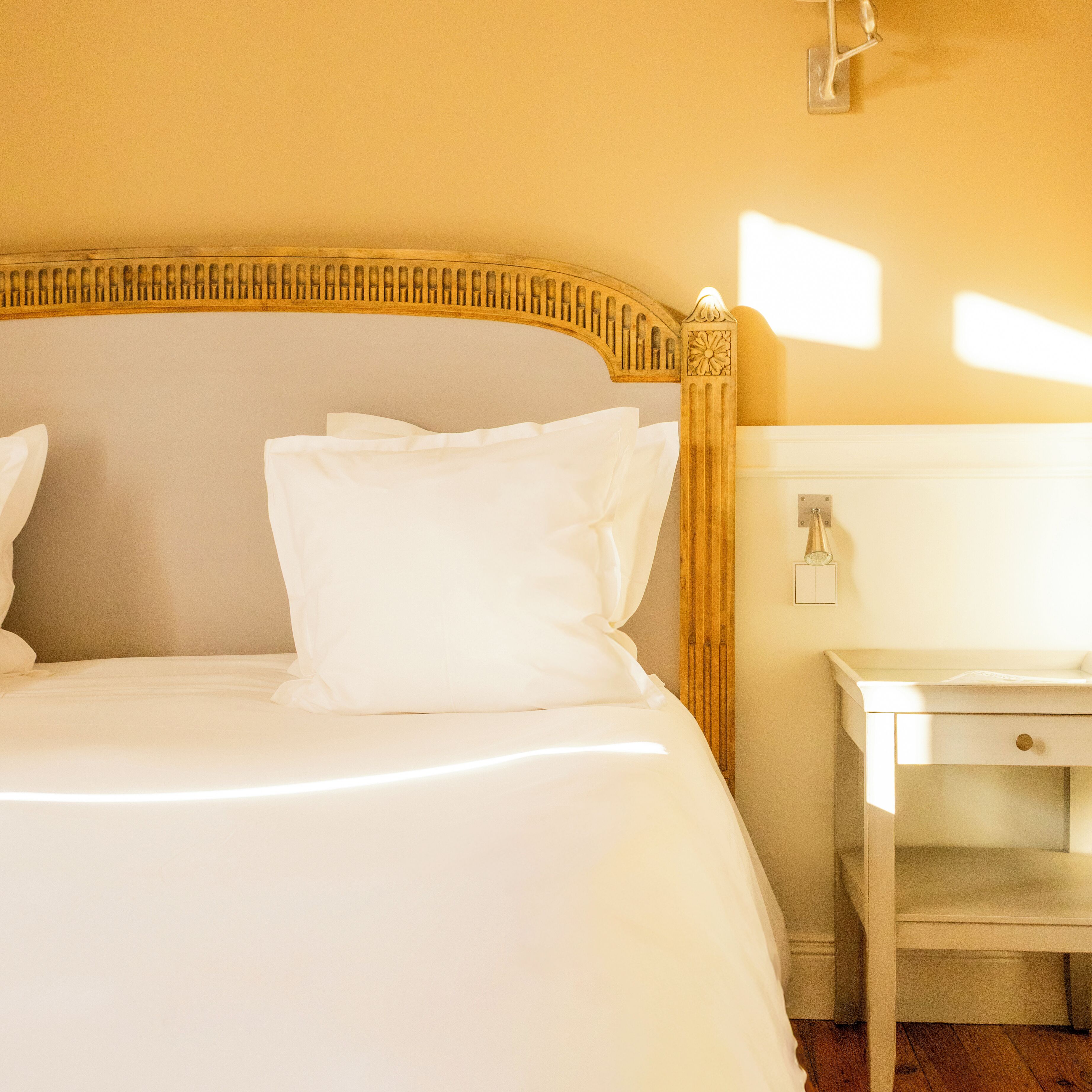 A neatly made bed with white linens and pillows, a wooden headboard with a decorative pattern, and a simple bedside table with a lamp.