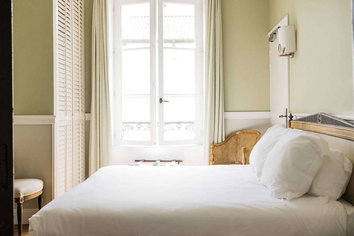 Ein Zimmer mit einem Bett, einem Sessel, einem Fenster mit Fensterläden und einer Wandleuchte.