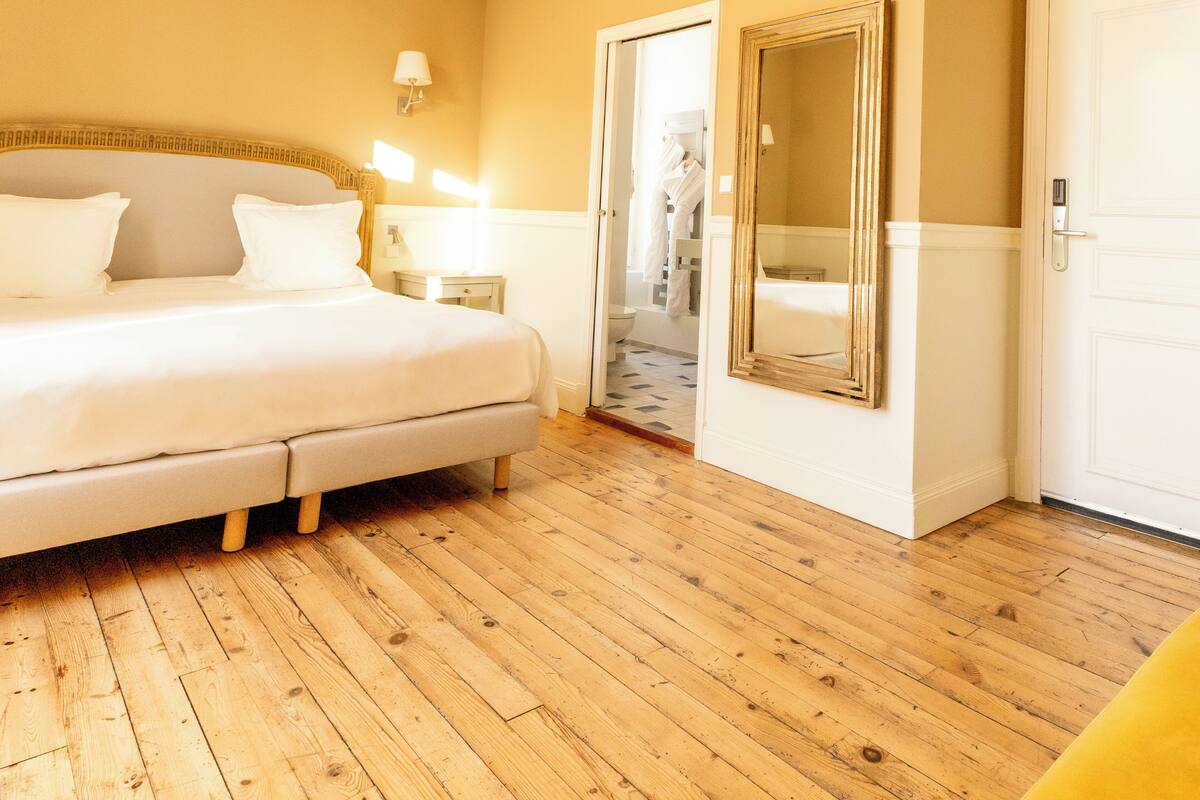Ein Hotelzimmer mit einem großen Bett, Holzboden, einem Spiegel und einer Tür zu einem Badezimmer.