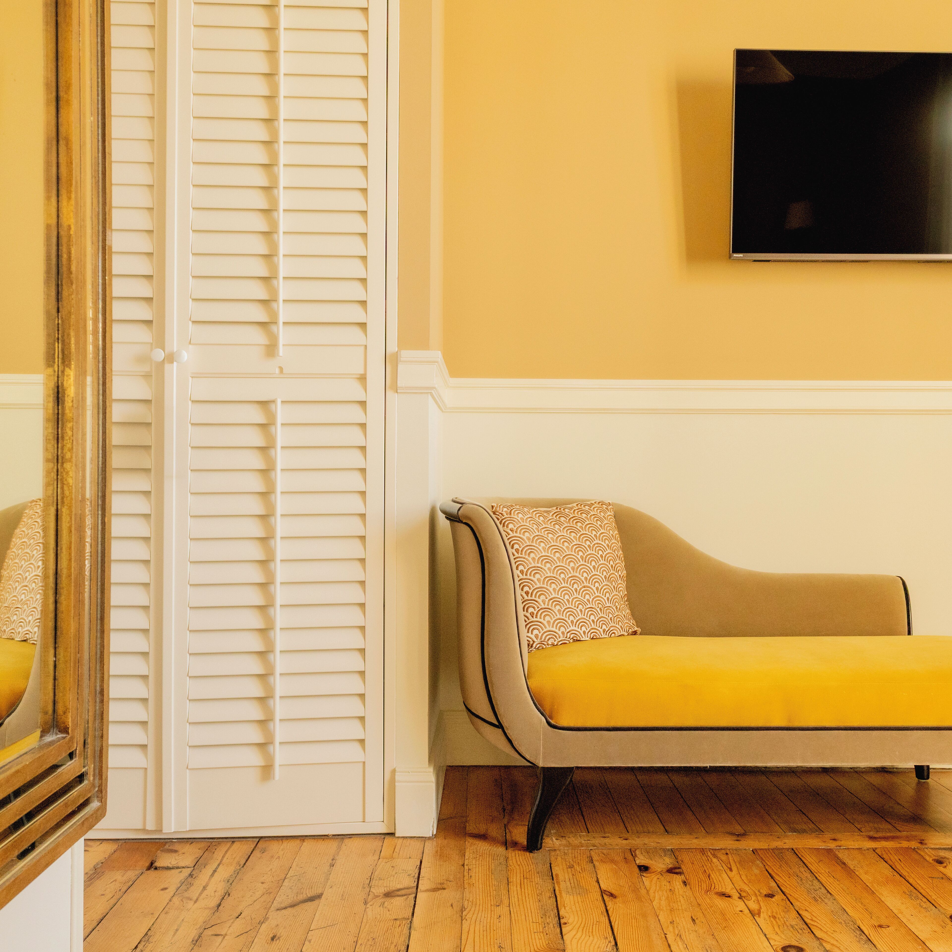 Una habitación con piso de madera, un sofá color beige con un cojín estampado, una puerta blanca con persianas y un televisor de pantalla plana montado en la pared.
