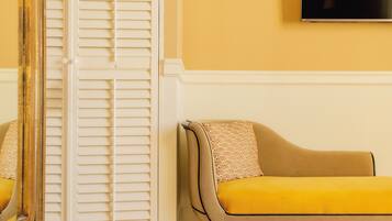 Una habitación con piso de madera, un sofá color beige con un cojín estampado, una puerta blanca con persianas y un televisor de pantalla plana montado en la pared.