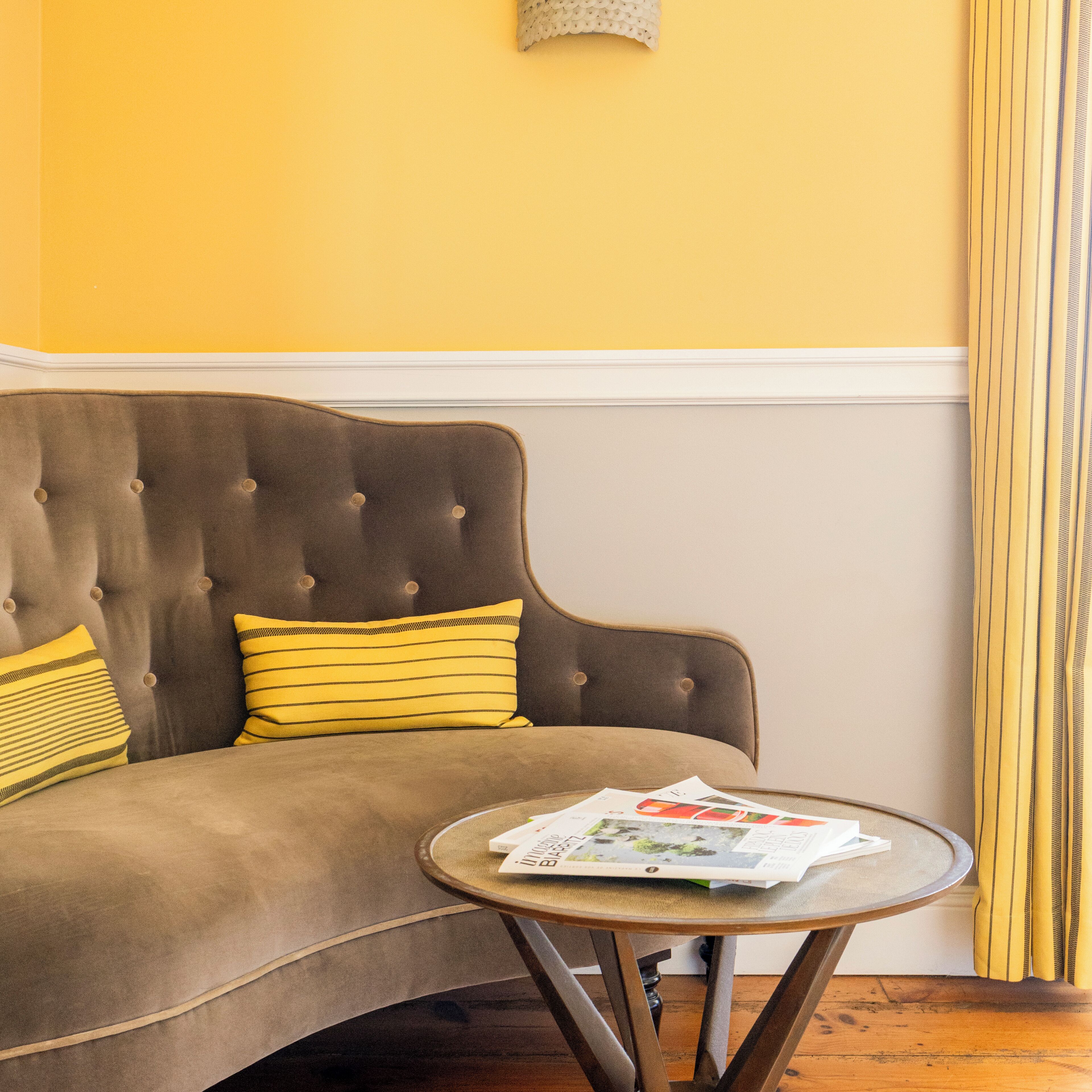 A brown sofa with yellow striped pillows, a round wooden table with magazines, and a yellow wall.