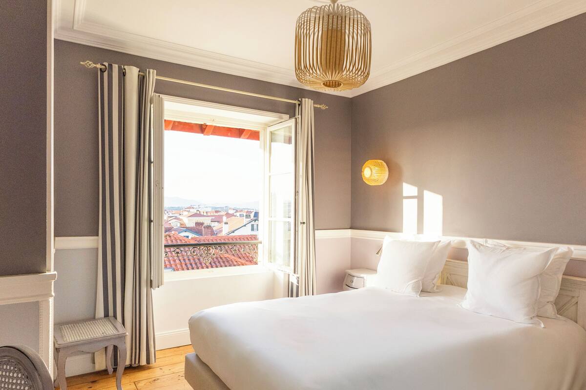 Ein Schlafzimmer mit einem großen Bett, einem Nachttisch, einem Sessel und Blick auf die Stadt durch ein offenes Fenster.