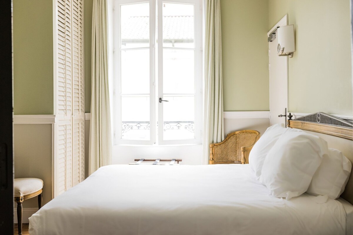 A room with a bed, a chair, a window with shutters, and a wall-mounted light fixture.