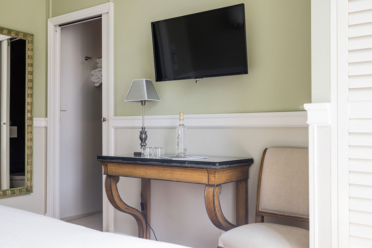 Ein Hotelzimmer mit an der Wand montiertem Fernseher, einem Nachttisch mit Lampe, einem Sessel und einem Spiegel.