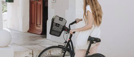 A woman standing next to a bicycle with a basket, in front of a hotel entrance with a red door and a sign with the letter 'H'.