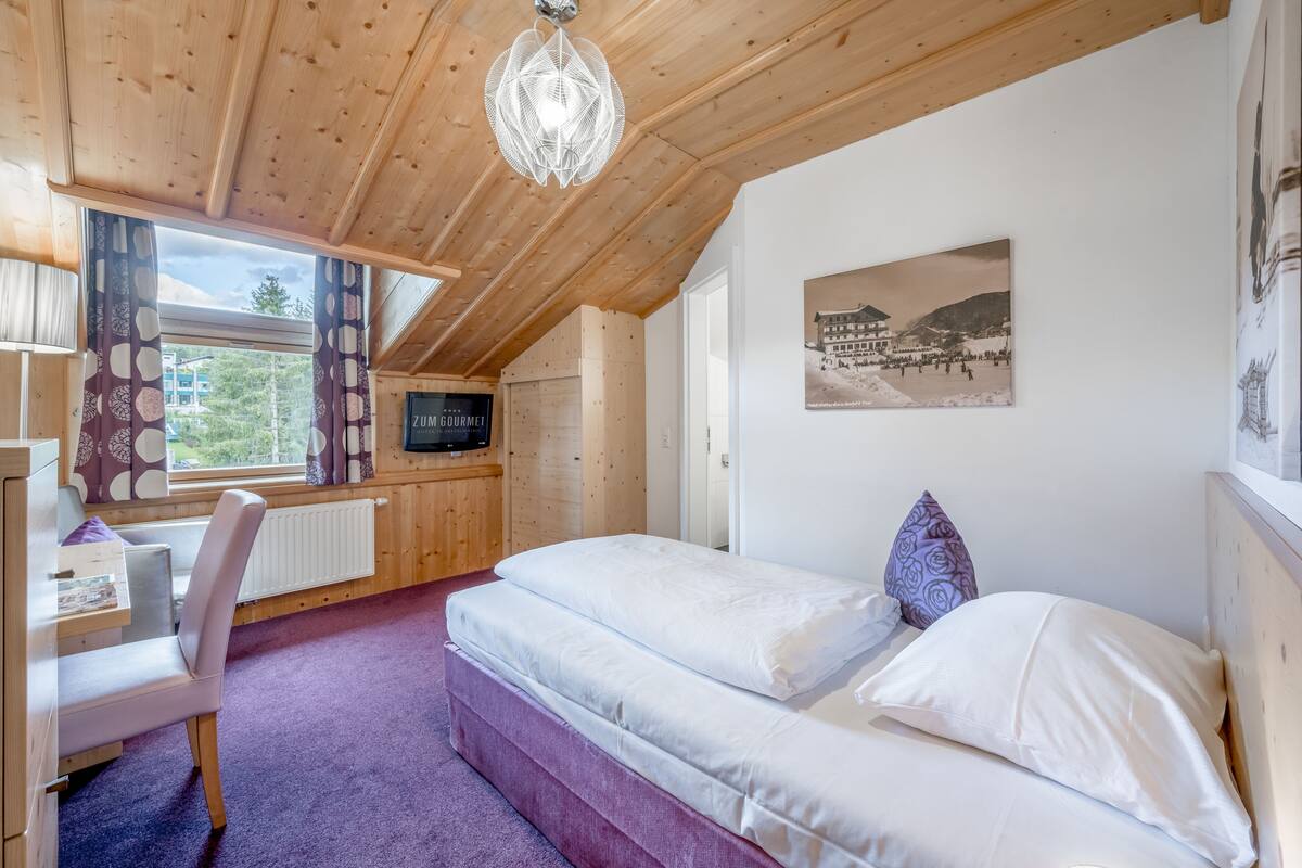 A cozy room with a wooden ceiling, a bed with white bedding, a desk with a chair, and a window with curtains.