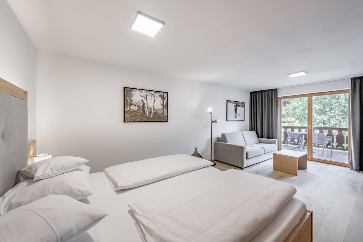 A modern hotel room with two beds, a sofa, a small table, and a balcony with outdoor furniture.