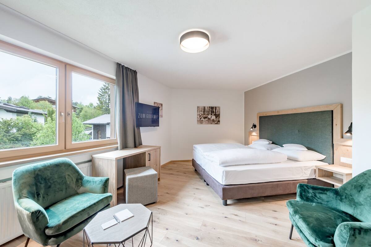 A hotel room with a bed, two green armchairs, a small table, a desk, and a window with a view of buildings and trees.