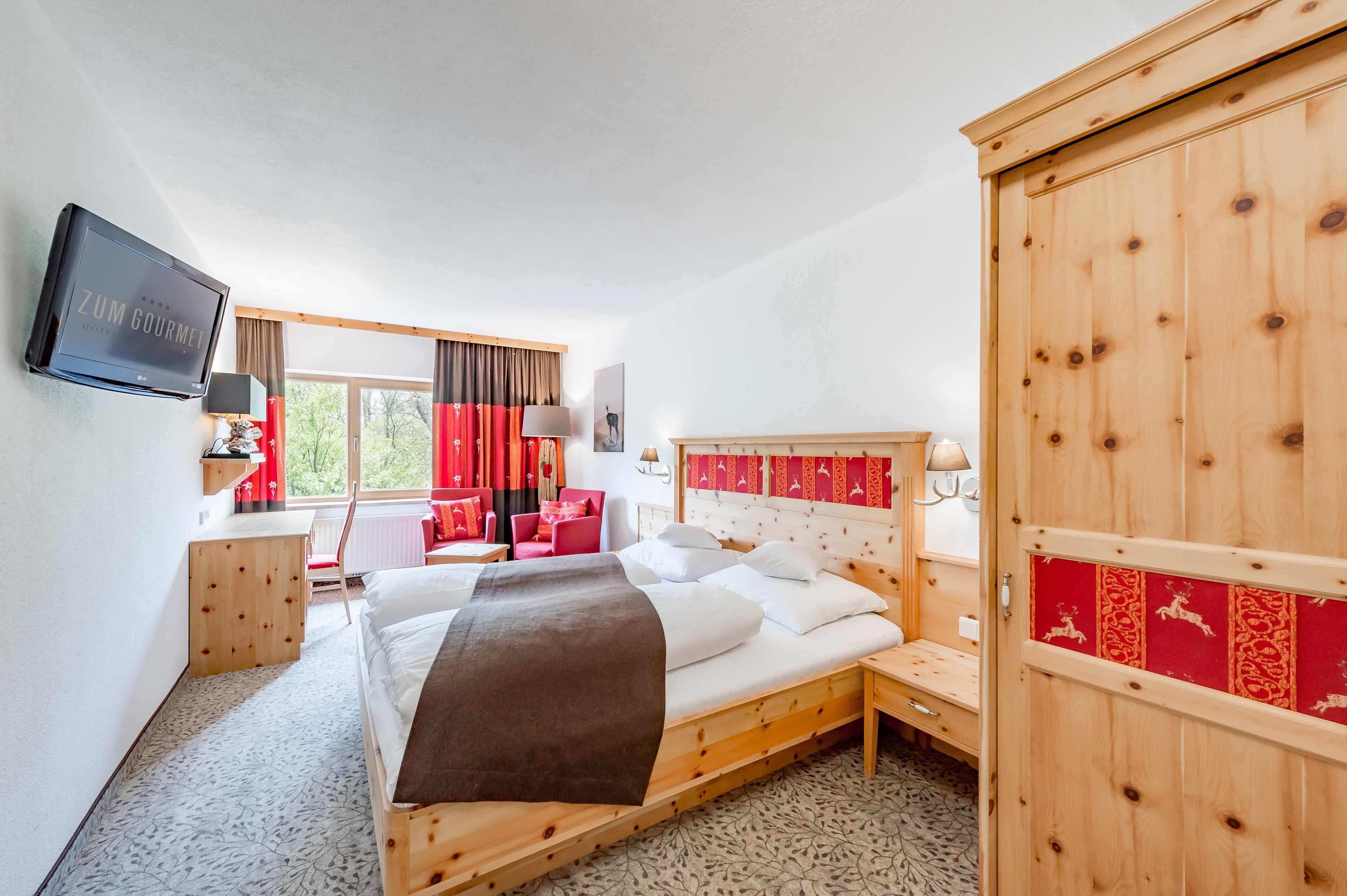 Quarto de hotel com cama de madeira, televisão fixada na parede, escrivaninha com cadeira e janela com cortinas.