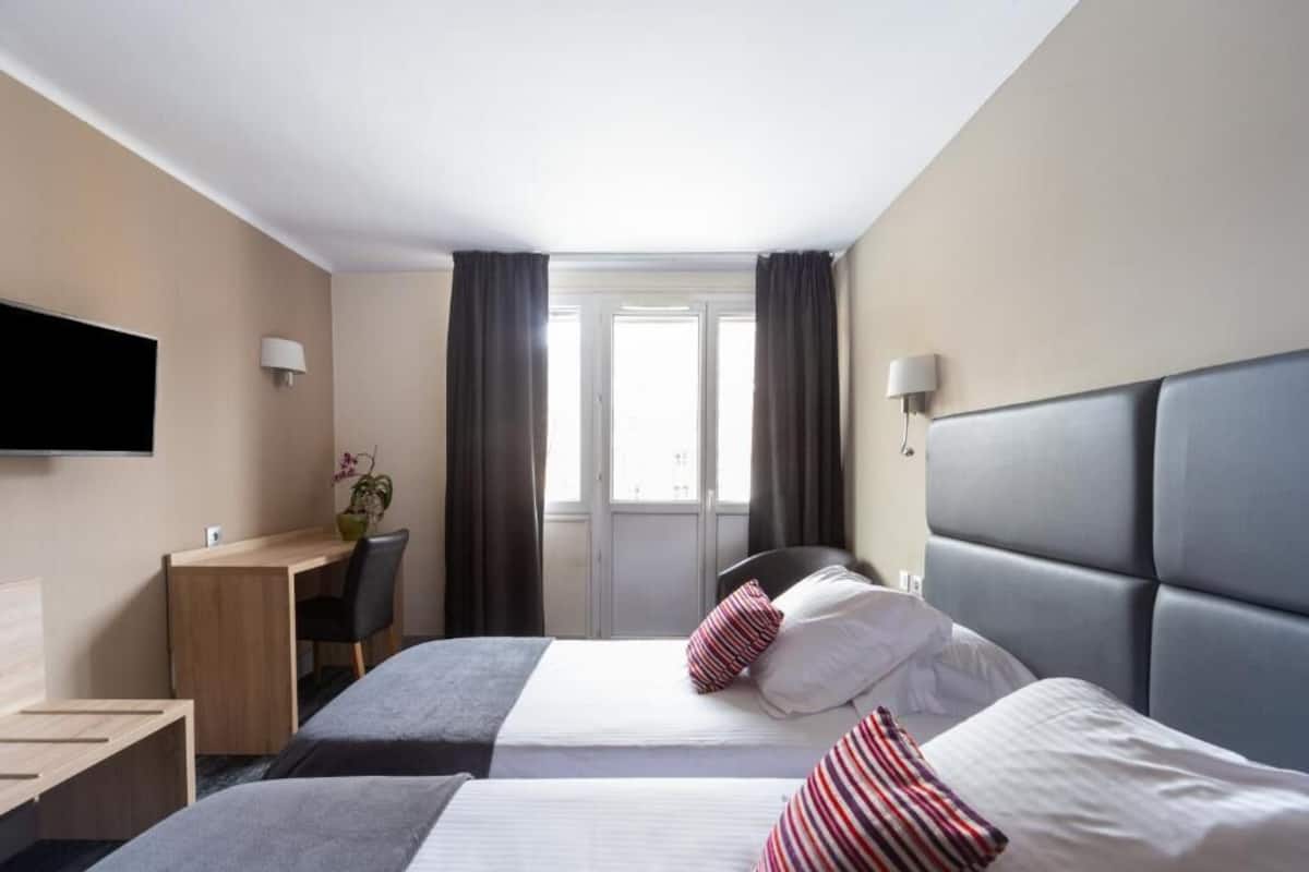 A hotel room with two beds, a desk, a chair, a TV, and a window with curtains.