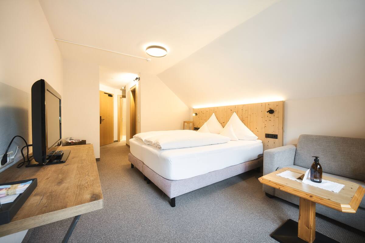 A modern hotel room with a bed, a sofa, a desk with a computer, and a wooden table.