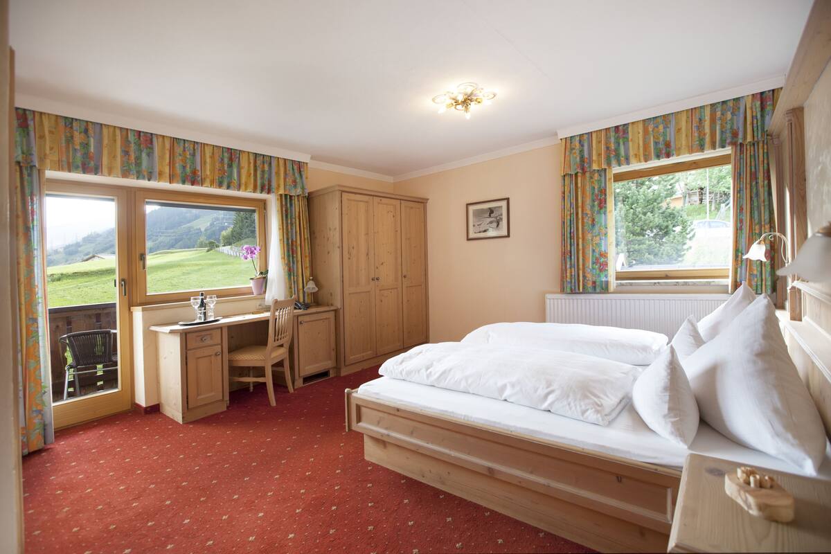 A bedroom with a bed, desk, chair, and large windows offering a view of the outdoors.