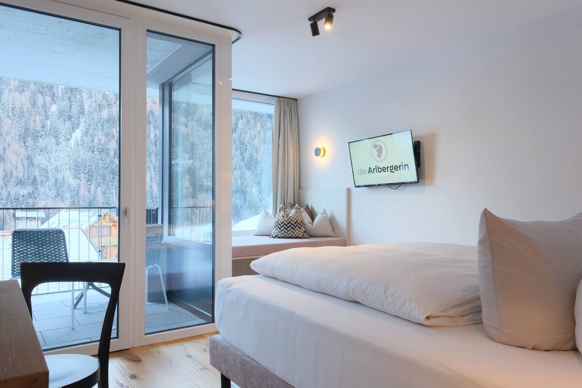 A hotel room with a large bed, a TV mounted on the wall, and a balcony with a view of snow-covered trees.