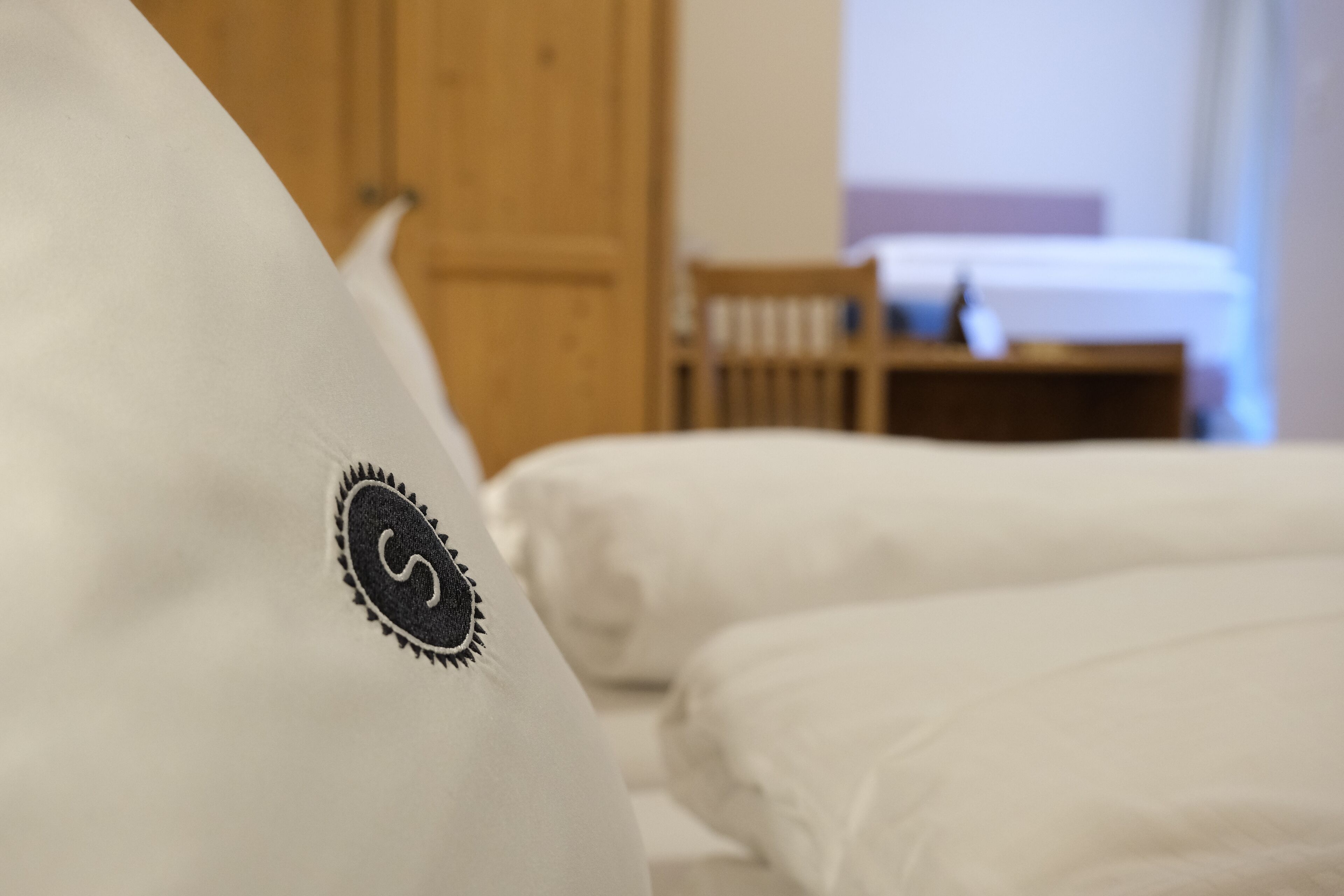 A close-up of a white pillow with a black embroidered logo. In the background, there is a wooden door and a bed with white bedding.