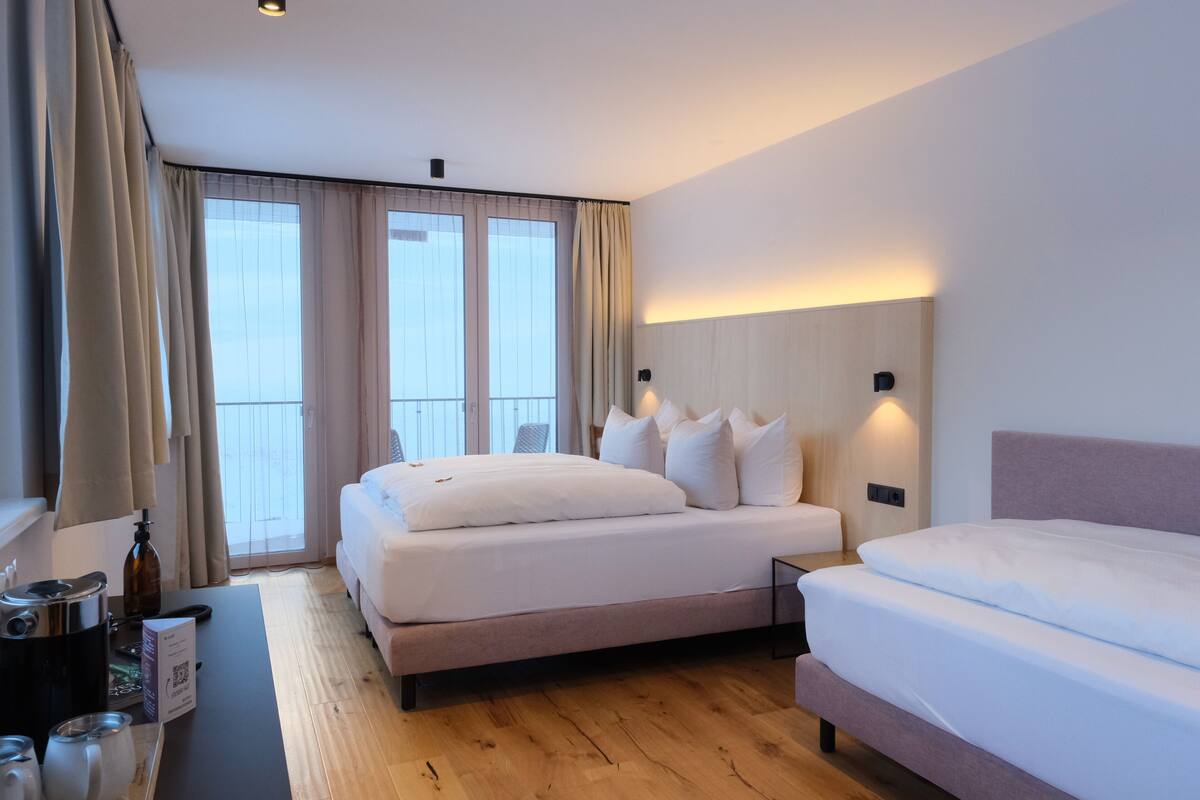 A modern hotel room with a large bed, a smaller bed, a desk with a coffee maker, and a view of the outdoors through large windows.