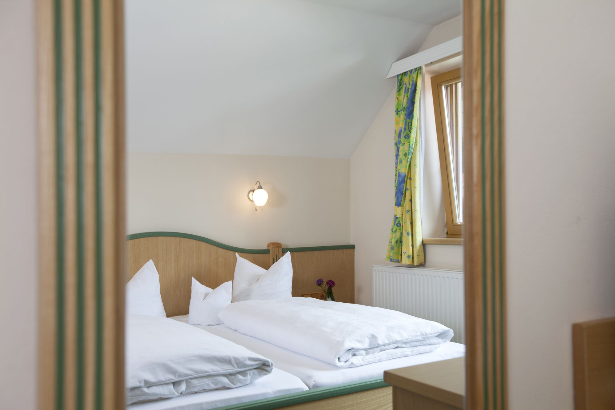 Uma cama de casal bem arrumada com roupa de cama branca, cabeceira de madeira, mesa de cabeceira e luminária fixada na parede.