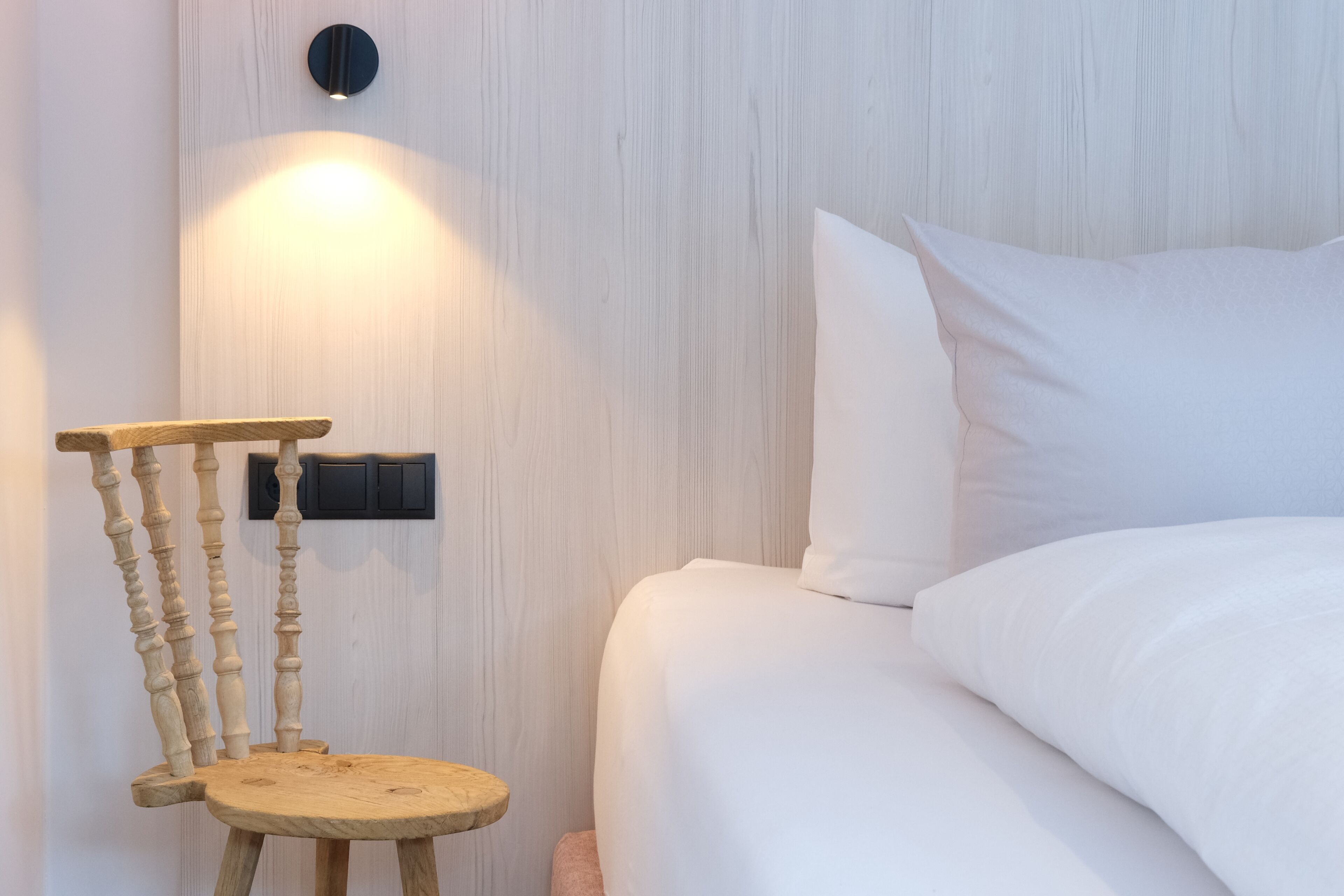 A bedroom with a wooden chair, a bedside table, and a wall-mounted light fixture.