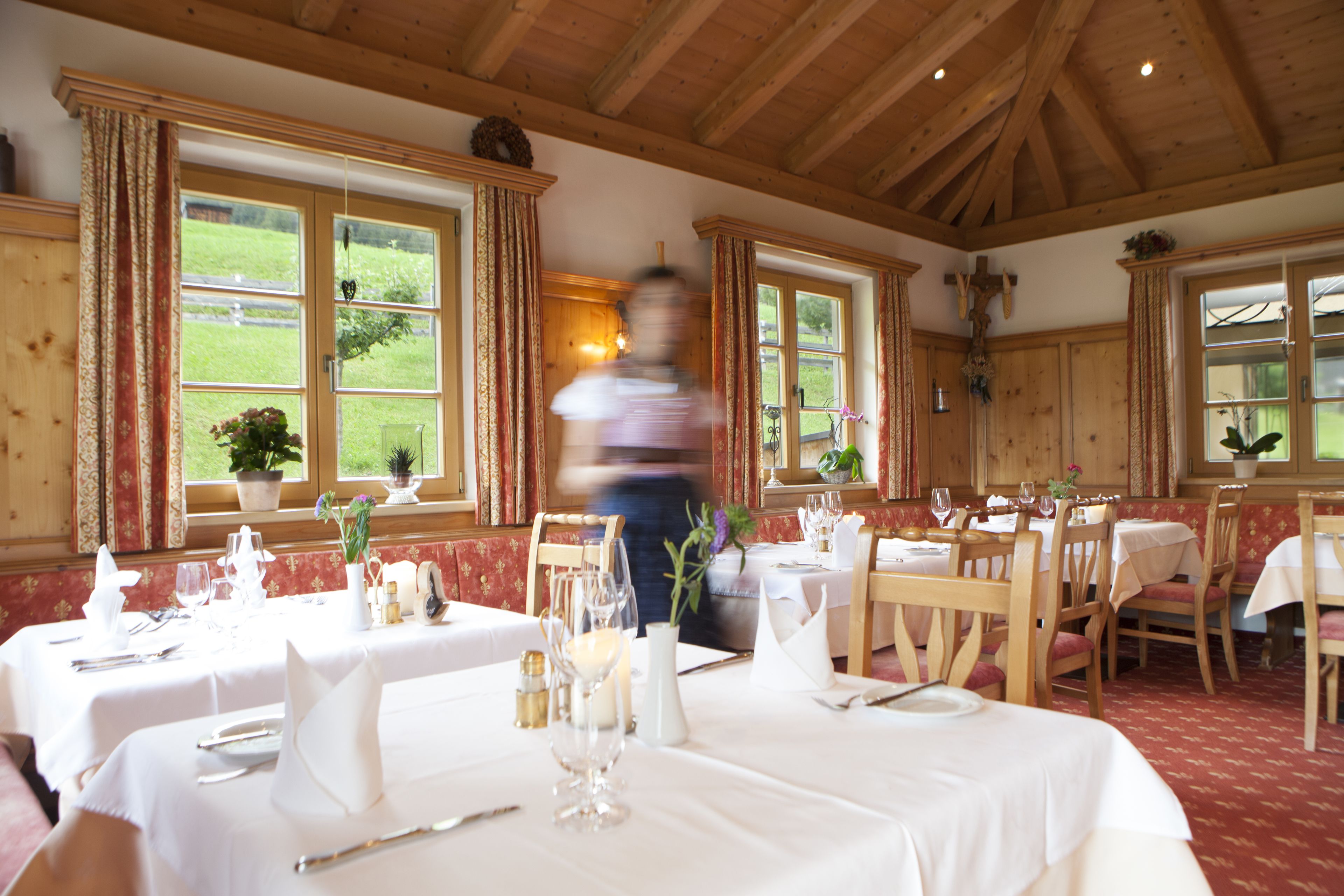 Un espace repas agrémenté de poutres en bois et de grandes fenêtres donnant sur un paysage verdoyant. Les tables sont dressées avec des nappes blanches et de beaux couverts.