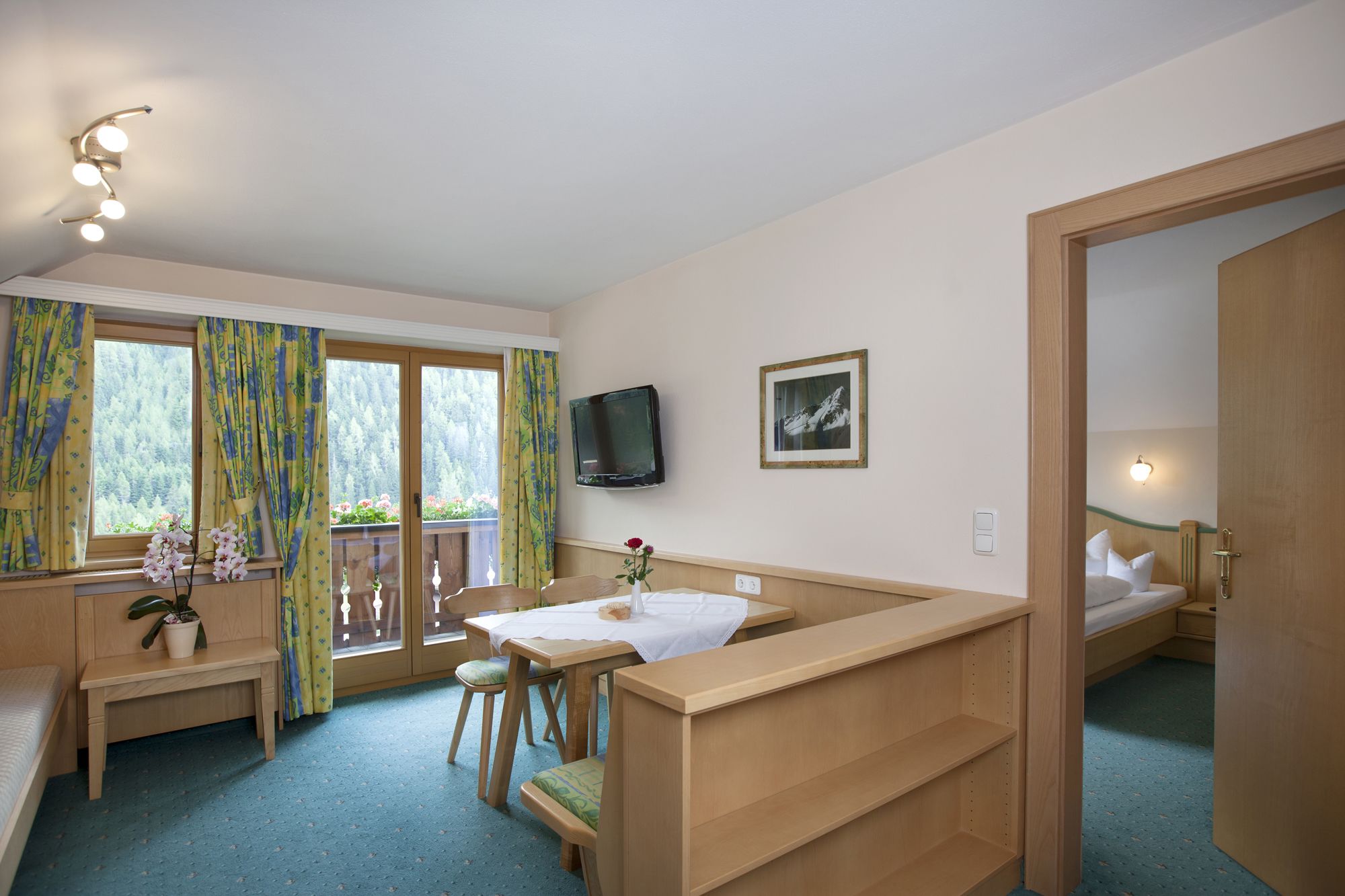 Een knus ingerichte kamer met een houten bureau, een stoel, een klein tafeltje met een vaas, een bed, een televisie en een raam met gordijnen.