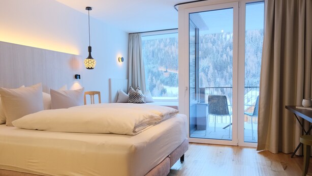 Ett modernt sovrum med en stor säng, ett sängbord, en stol och utsikt över ett snötäckt landskap genom en skjutdörr i glas.