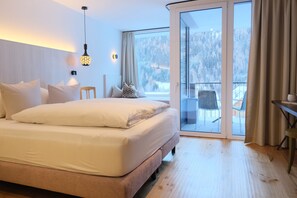 A modern bedroom with a large bed, a bedside table, a chair, and a view of a snowy landscape through a sliding glass door. - Hotel die Arlbergerin - Adults Friendly (Sankt Anton am Arlberg)