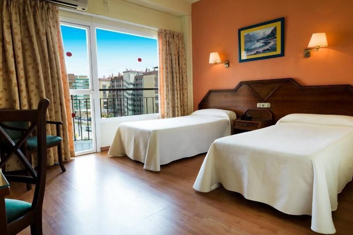 Una habitación de hotel con dos camas, una silla, una mesa, una mesita de noche y un cuadro en la pared.