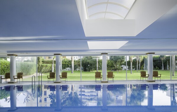 A modern indoor swimming pool with clear glass walls, allowing views of a well-maintained lawn and trees.