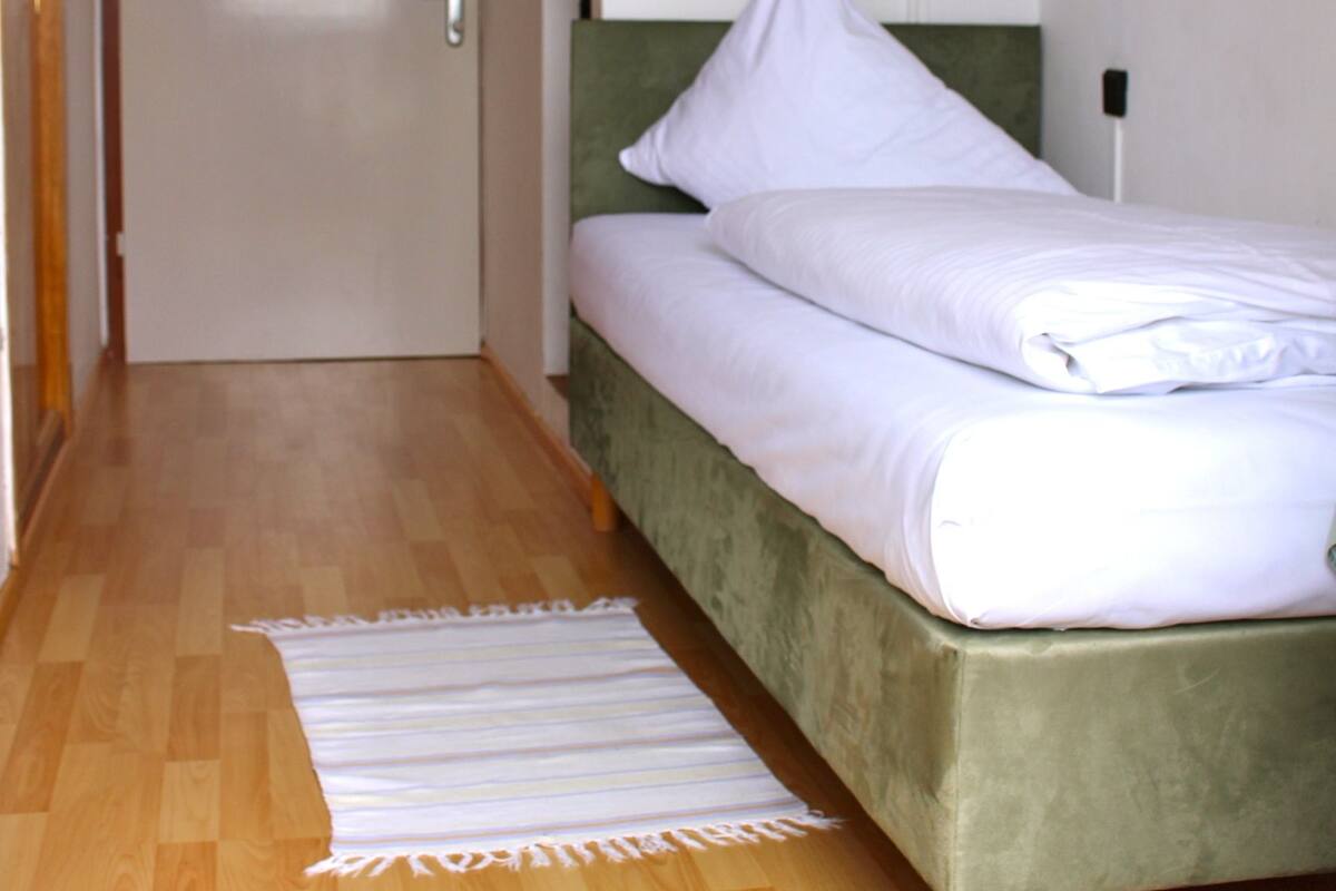 A bedroom with a green bed, white bedding, a wooden floor, and a hanging light fixture.