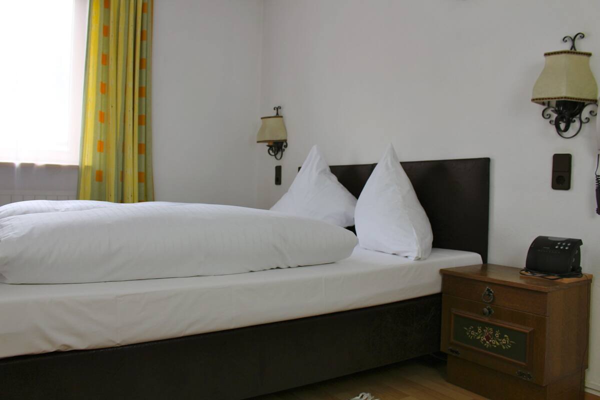 A neatly made bed with white linens, a wooden nightstand with a lamp, and a wall-mounted telephone.