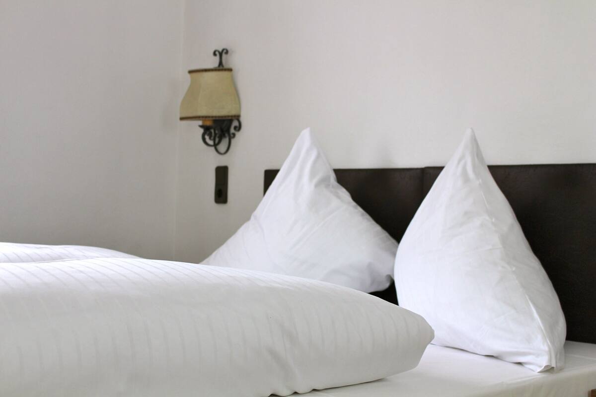 A neatly made bed with white linens and pillows, a dark headboard, and a wall-mounted lamp.