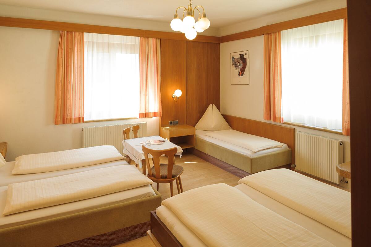 A hotel room with four beds arranged in a U-shape, a wooden-framed desk with a chair, a table, and a wall-mounted lamp.