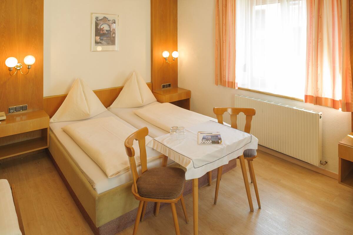 A hotel room with a double bed, two bedside tables, a small table with a chair, and a window with curtains.