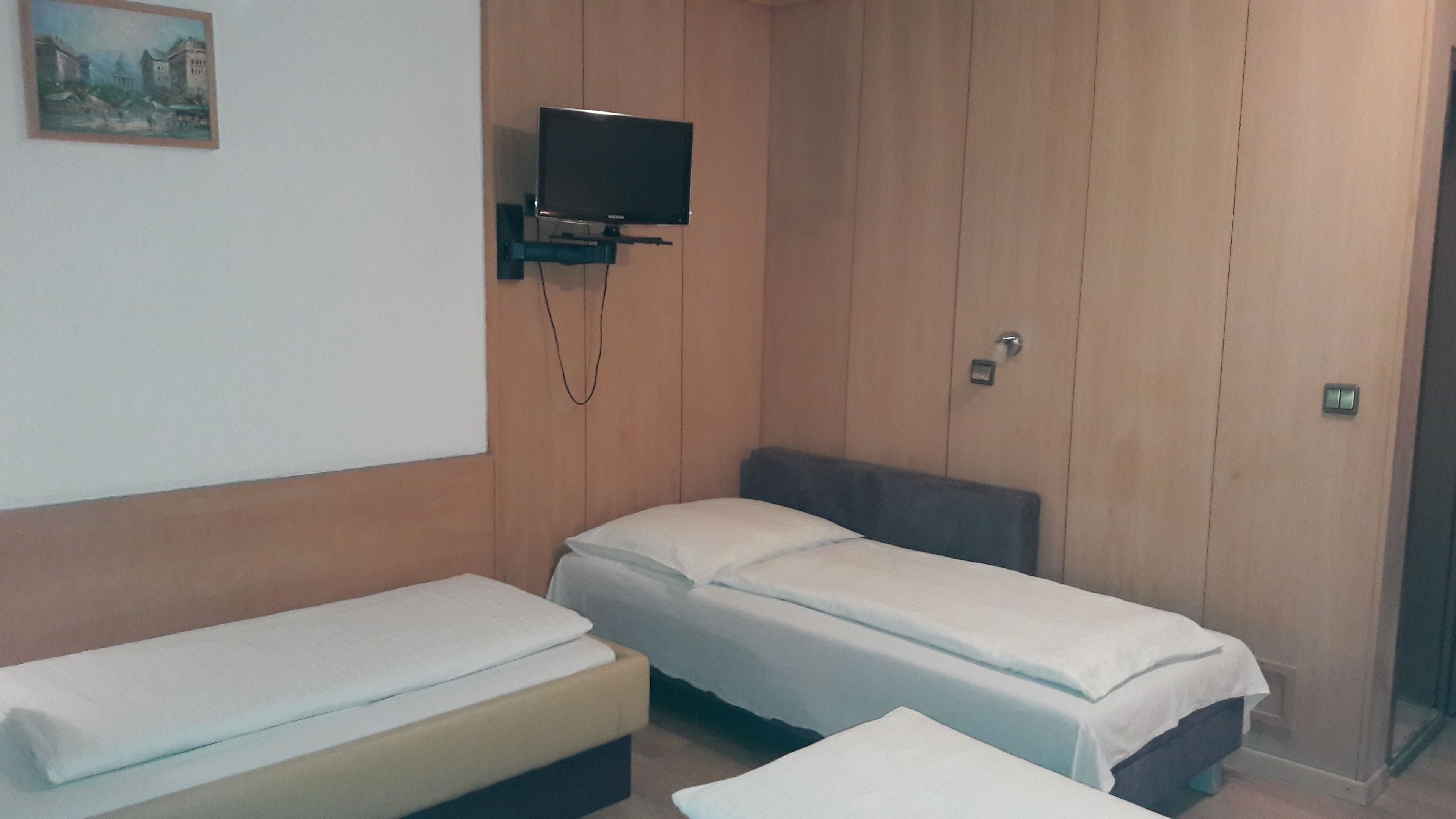 A small, single-room hotel room with a bed, a television mounted on the wall, and a wooden panel wall.
