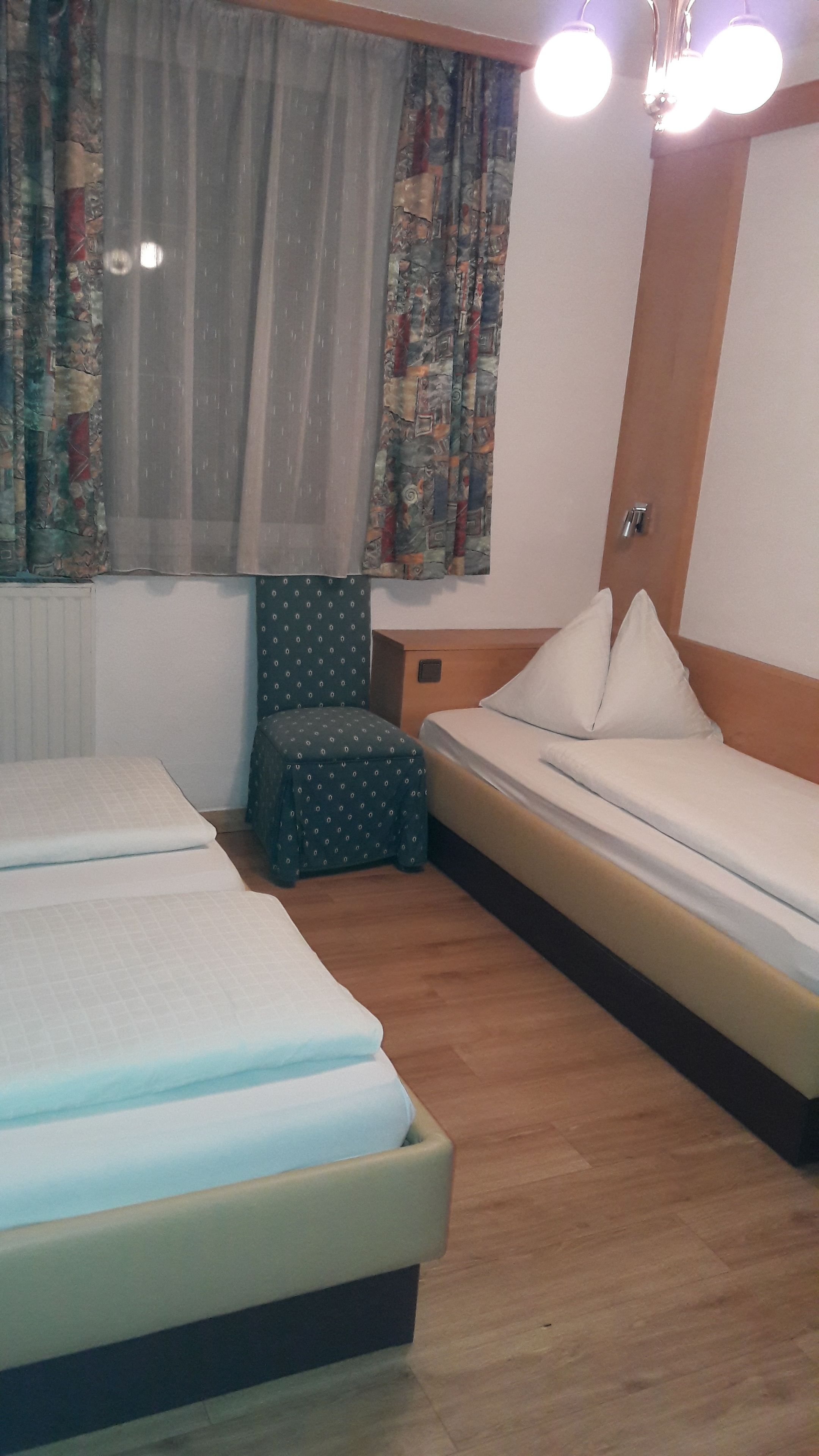 Habitación de hotel con dos camas, un taburete pequeño, una ventana con cortinas estampadas y un cabecero de madera.