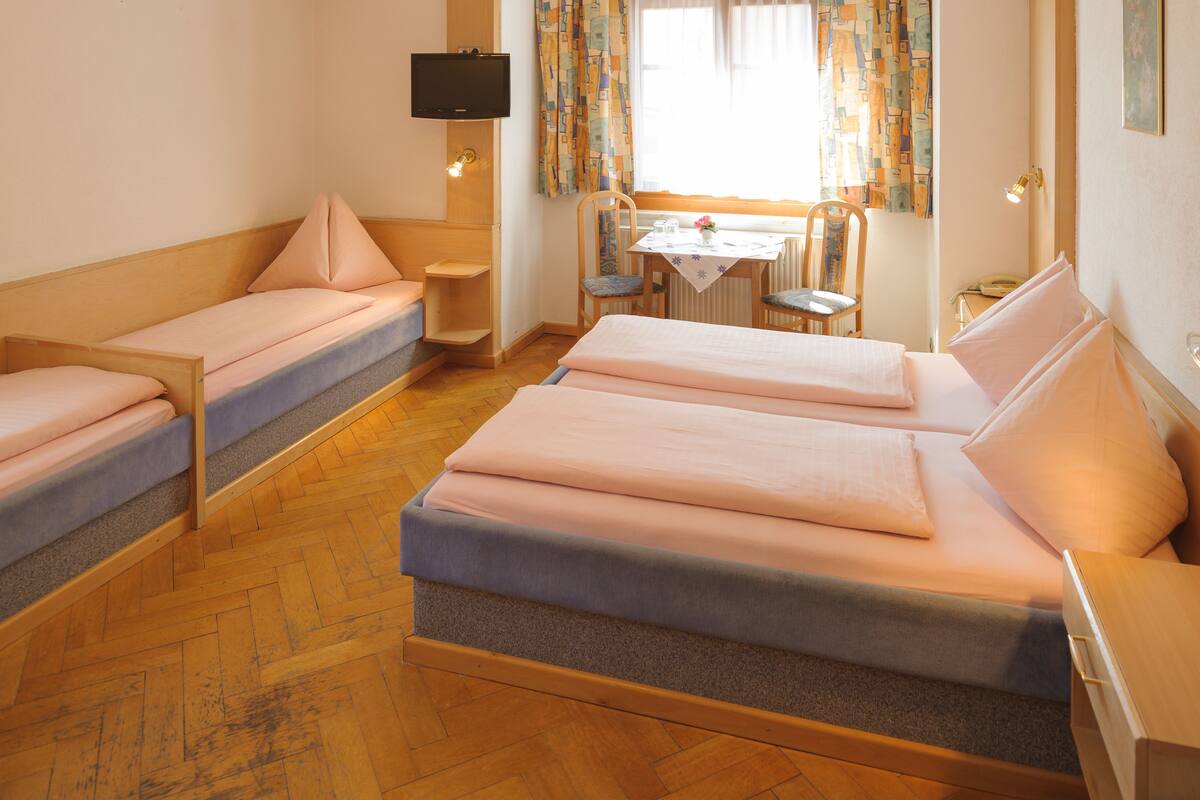 A room with two single beds, a small table with chairs, a television, and a window with curtains.