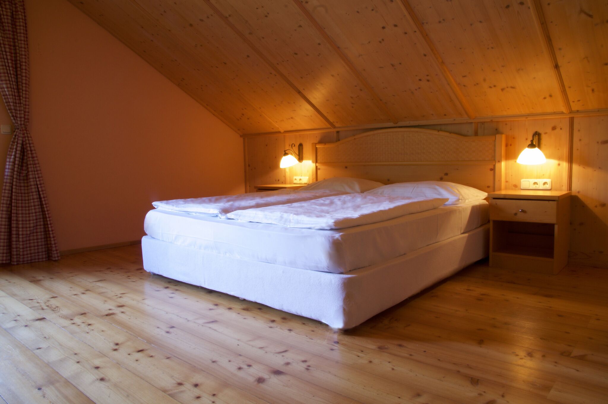 Un dormitorio con techo de madera, una cama con ropa de cama blanca, dos mesitas de noche con lámparas y suelo de madera.